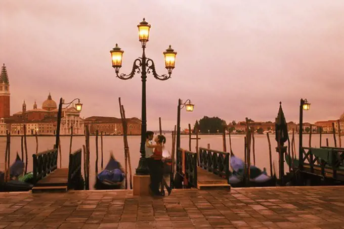 Couple embracing against lamp post on waterside walkway near St Marks Square in Venice, Italy