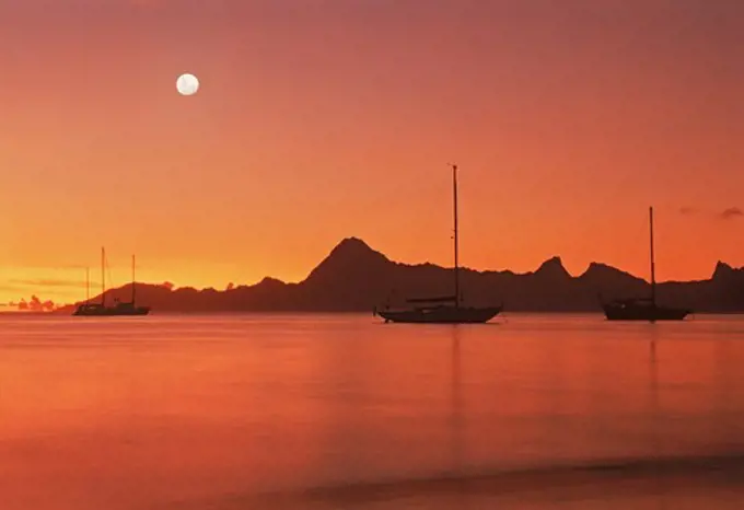 Full moon over distant Moorea from island of  Tahiti with sailboats anchored offshore at sunset