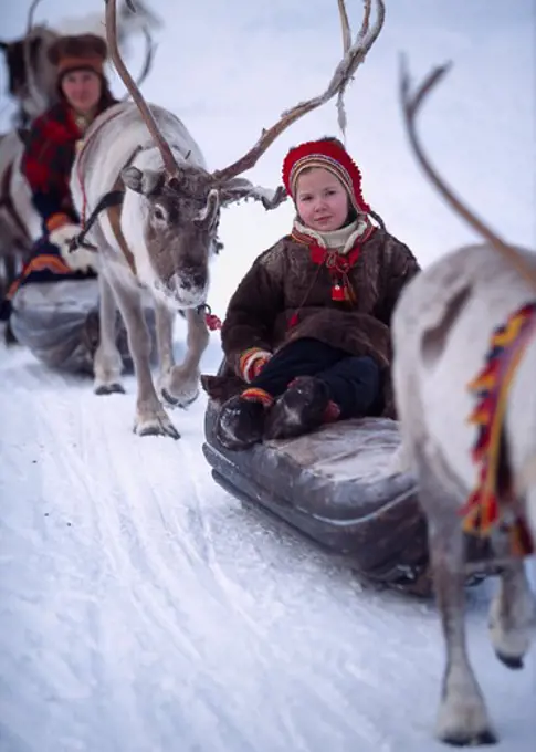 Reindeers pulling two girls sitting on the sleds on a snow covered landscape, Jokkmokk, Lapland, Sweden