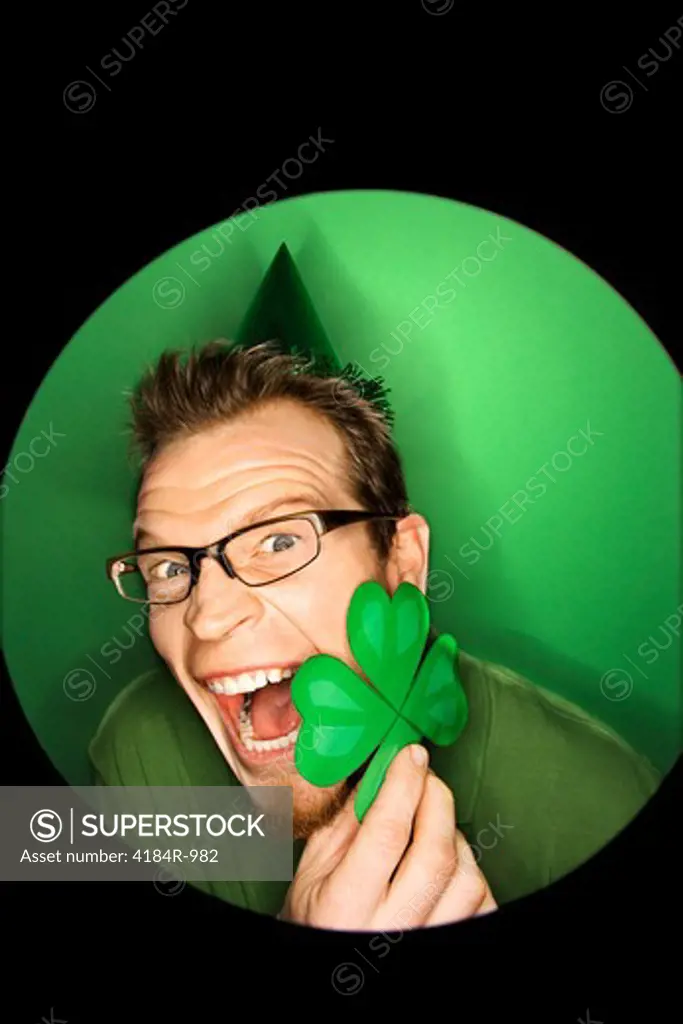 Vignette of excited adult Caucasian man on green background wearing Saint Patricks Day hat and holding shamrock.