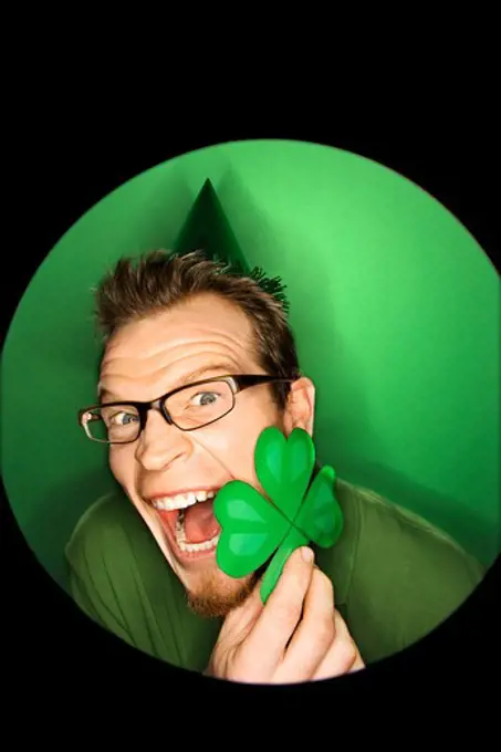 Vignette of excited adult Caucasian man on green background wearing Saint Patricks Day hat and holding shamrock.