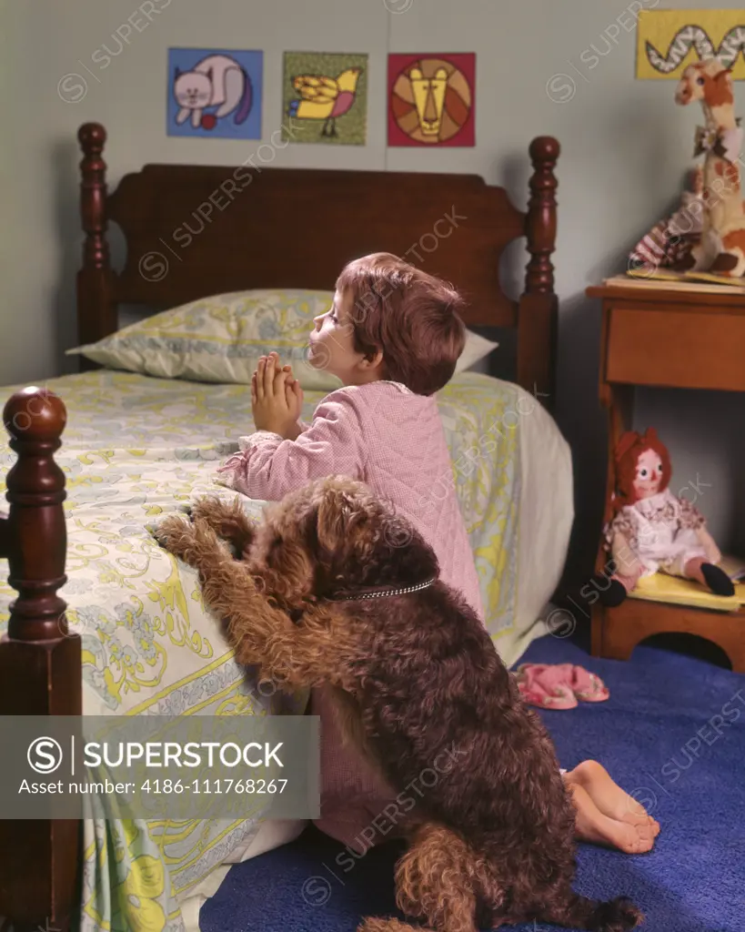 1970s 1980s LITTLE RED HAIR GIRL KNEELING IN BEDROOM BESIDE BED SAYING PRAYERS WITH PET WIRE HAIR TERRIER DOG ALONGSIDE