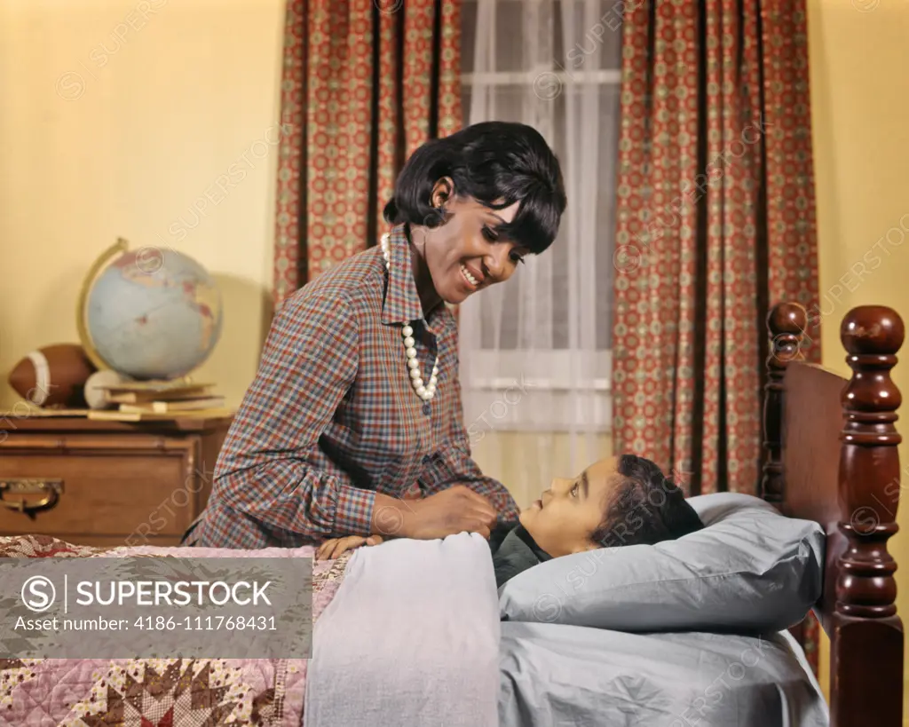 1970s SMILING AFRICAN AMERICAN WOMAN MOTHER TUCKING GIRL DAUGHTER INTO BED AT NIGHT