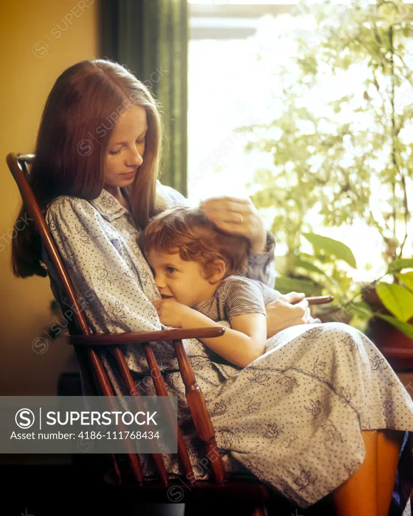 1970s WOMAN SITTING IN ROCKING CHAIR COMFORTING BOY TODDLER STANDING NEXT TO HER BY HOUSEPLANT FILLED WINDOW