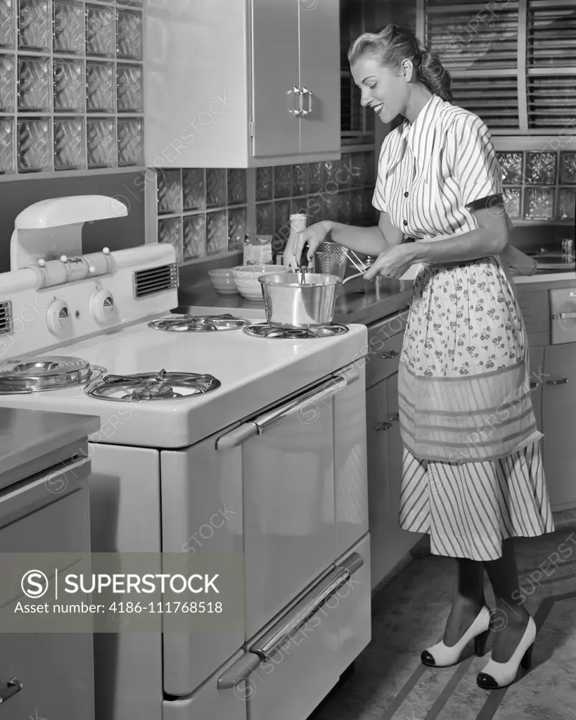 1950s SMILING BLOND HOUSEWIFE WEARING DRESS APRON TWO TONE HIGH HEEL SHOES COOKING ON MID-CENTURY MODERN ELECTRIC KITCHEN RANGE