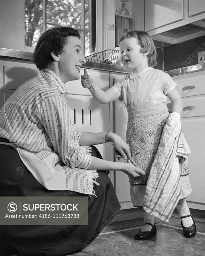 1950s LITTLE GIRL AND MOTHER IN KITCHEN BOTH WEARING APRONS GIRL HOLDING DISH TOWEL GESTURING TO MOTHER