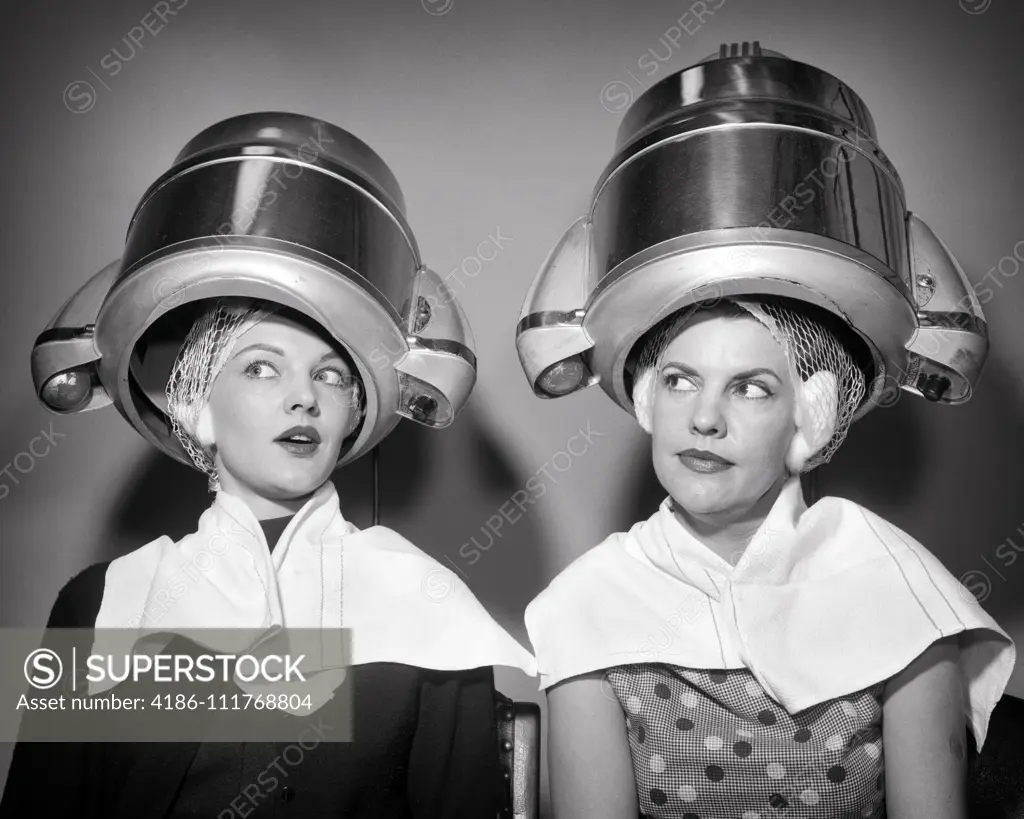 1950s TWO WOMEN SITTING UNDER BEAUTY SALON HAIR DRYERS WEARING HAIRNETS TOWELS TALKING GOSSIP