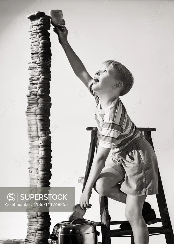 1950s EAGER BOY STANDING ON STEP LADDER REACHING THE TOP BUILDING A TOWER OF TOASTED BREAD SLICES