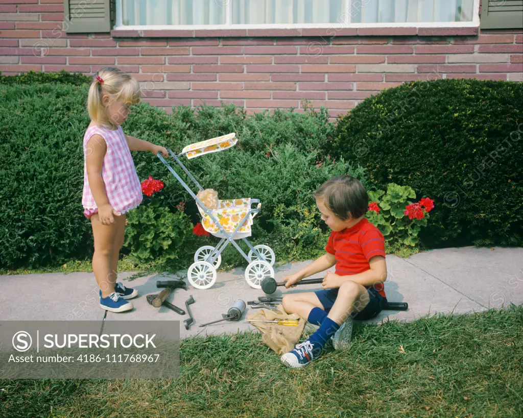 1970s LITTLE GIRL PUSHING TOY BABY CARRIAGE AND BOY PLAYING WITH TOOLS ON SIDEWALK GENDER SPECIFIC ROLE PLAY