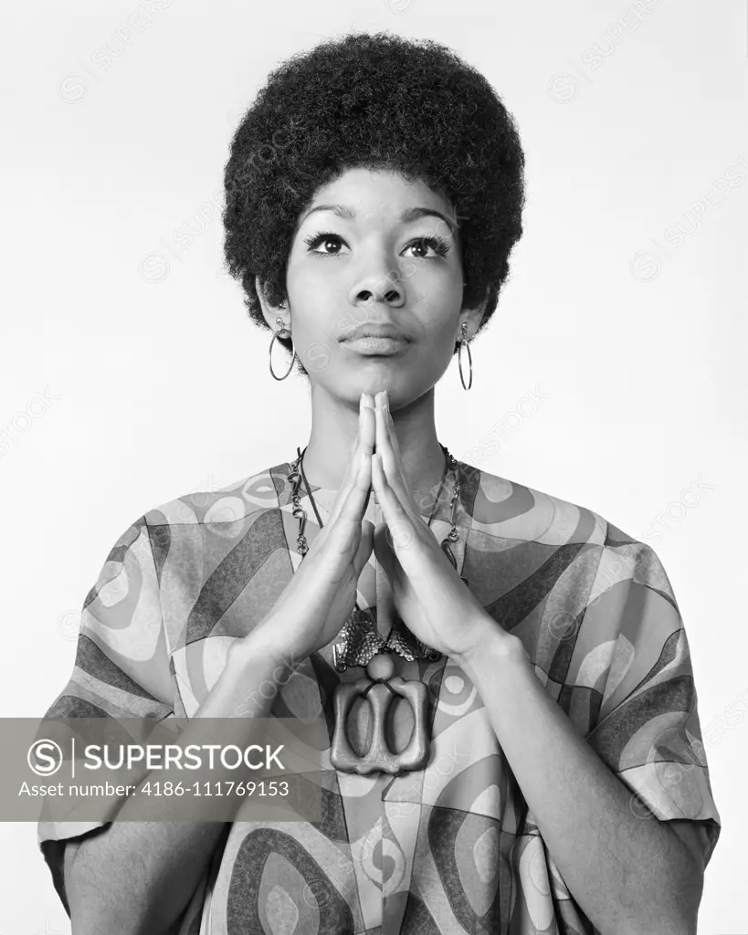 1960s AFRICAN AMERICAN WOMAN AFRO HAIRDO LOOKING AT CAMERA ARMS FOLDED IN PRAYER MEDITATION WEARING HOOP EARRINGS PRINT BLOUSE