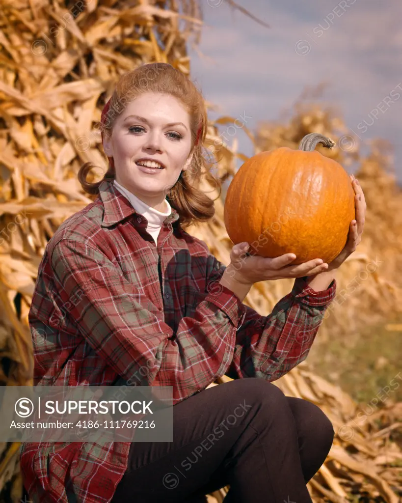 1960s SMILING RED HAIR WOMAN WEARING A PLAID SHIRT SITTING BY CORN SHOCK LOOKING AT CAMERA HOLDING UP PUMPKIN