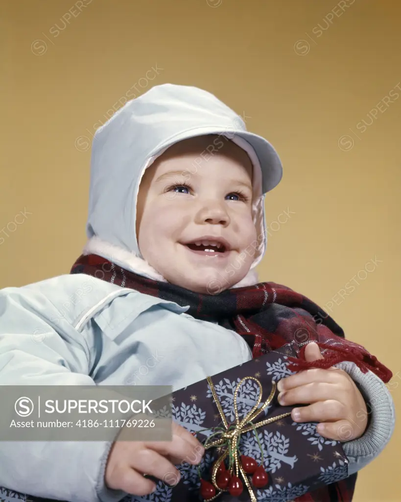 1960s SMILING TODDLER BOY WEARING WINTER SNOWSUIT HOLDING WRAPPED CHRISTMAS GIFT