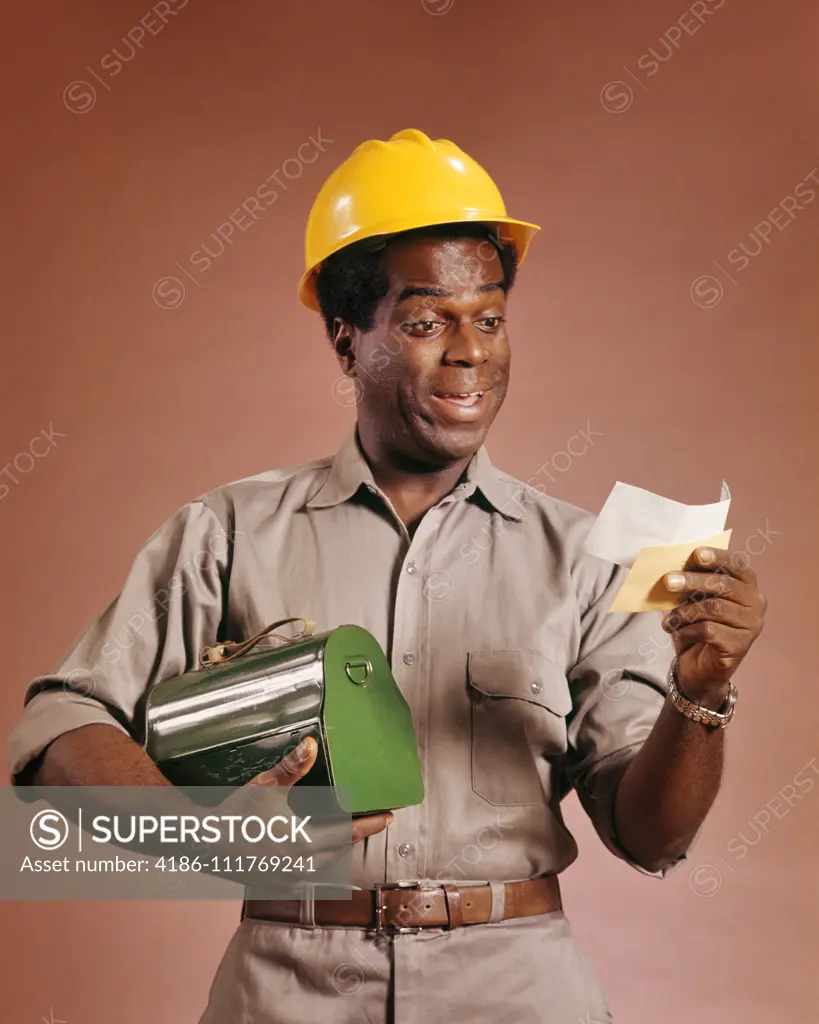 1970s AFRICAN AMERICAN WORKMAN HOLDING LUNCHBOX AND PAYCHECK SMILING WEARING A YELLOW HARD HAT