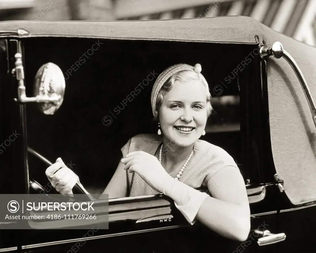 1920S 1930s SMILING WOMAN DRIVING CAR LOOKING OUT WINDOW WEARING GLOVES PEARL NECKLACE ELEGANTLY DRESSED