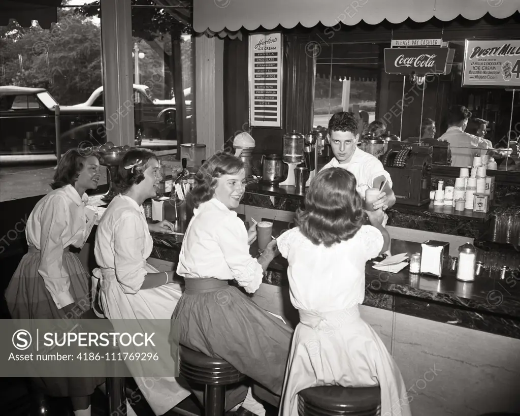 1950s FOUR TEENAGE GIRLS SEATED AND SERVED ICE CREAM SODAS AT COUNTER OF SUBURBAN DRUG STORE BY BOY SODA JERK BRONXVILLE NY USA