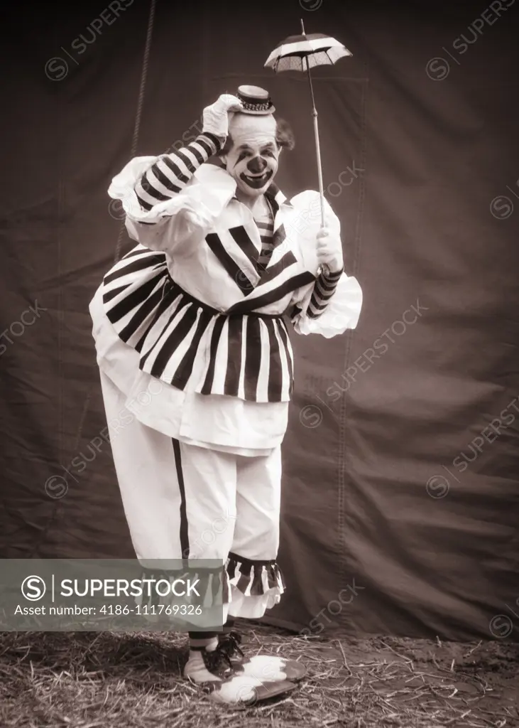 1930s BIG CLOWN UNDER TINY UMBRELLA TIPPING HIS SMALL HAT LOOKING AT CAMERA WEARING CLOWN SHOES OVERSTUFFED STRIPED COSTUME