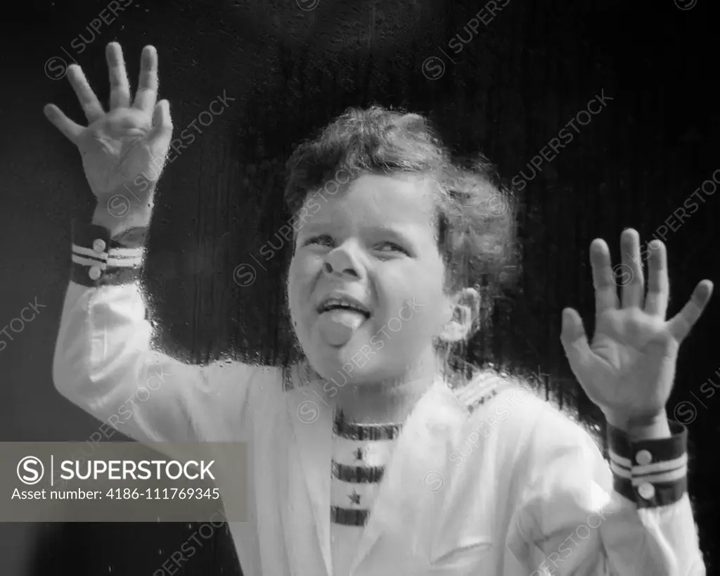 1930s ODD LAUGHING CURLY HAIR BOY WEARING NAUTICAL STYLE SHIRT PLACING HANDS AND STICKING TONGUE ON RAIN WET WINDOW GLASS PANE