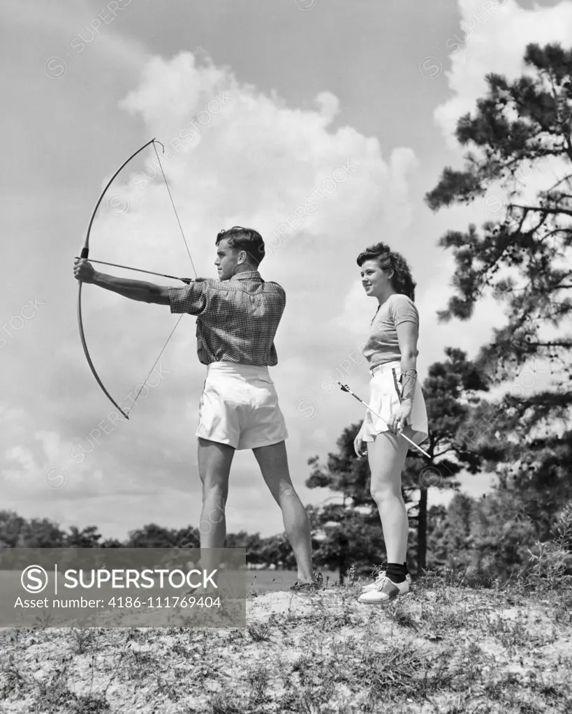 1930s TEENAGE COUPLE PRACTICING ARCHERY SHOOTING BOW AND ARROW