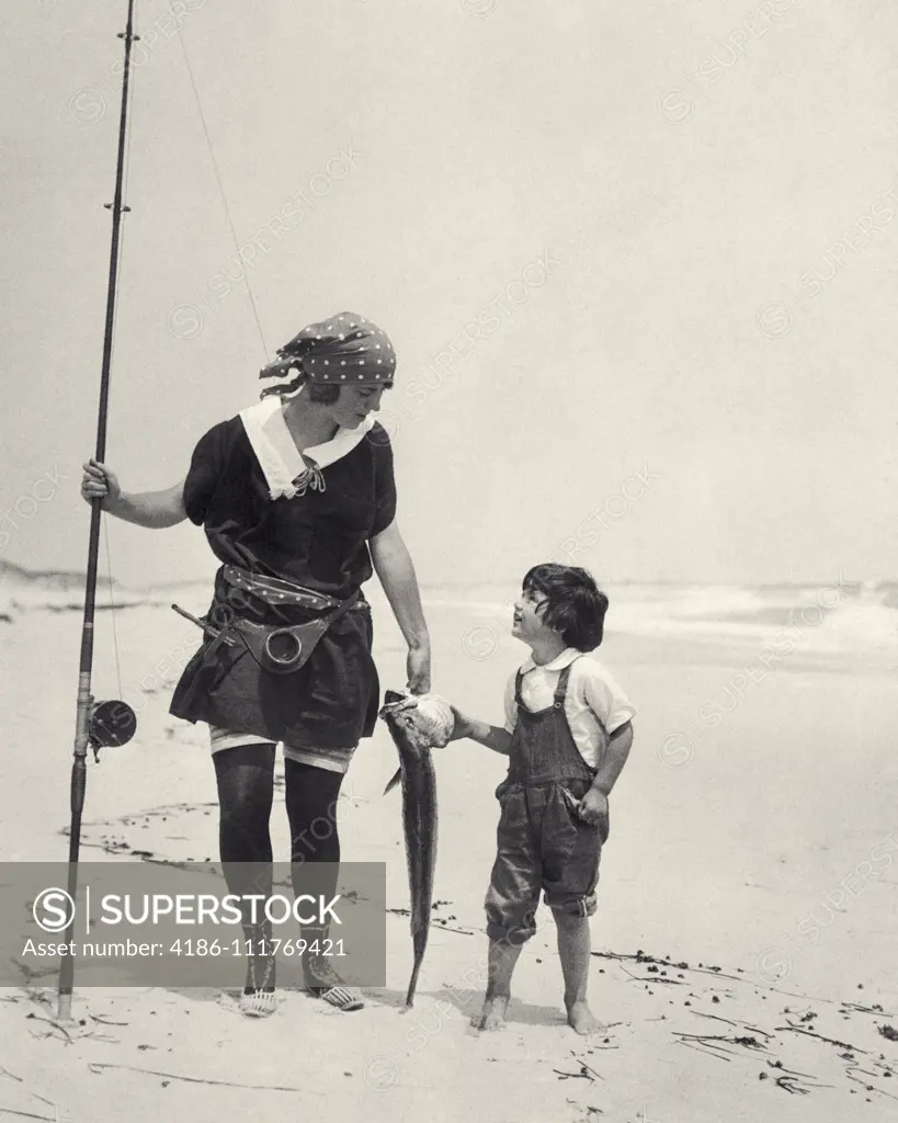 1920s WOMAN MOTHER HOLDING SURF CASTING FISHING POLE CARRYING FISH CATCH WITH BOY SON WALKING ON  OCEAN SEASHORE BEACH SAND