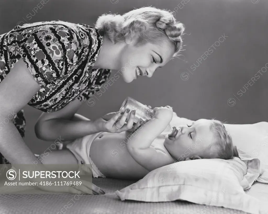 1930s SMILING BLOND MOTHER BENDING OVER FEEDING MILK BOTTLE TO BABY LYING  ON BACK - SuperStock