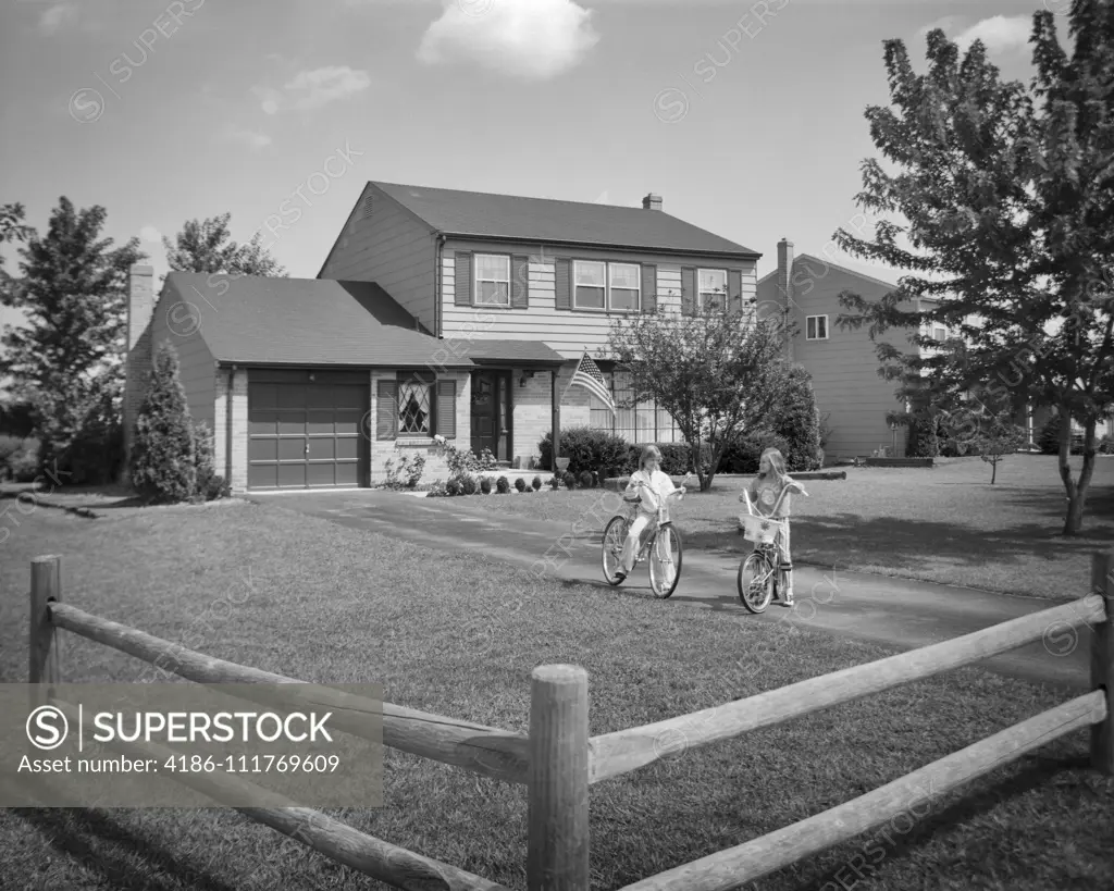 1970s TWO YOUNG GIRLS SISTERS RIDING BICYCLES IN YARD OF SUBURBAN HOUSE