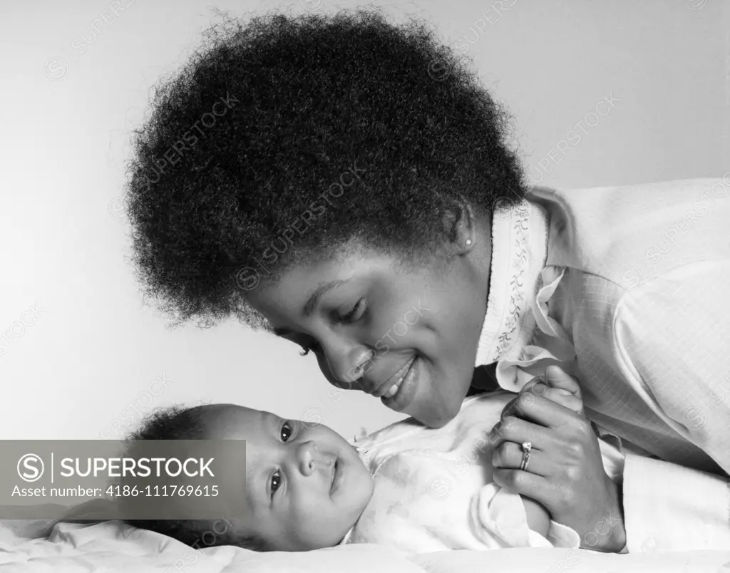 1970s SMILING AFRICAN AMERICAN MOTHER HOLDING HAND AND LOOKING AT HAPPY BABY DAUGHTER LYING ON HER BACK