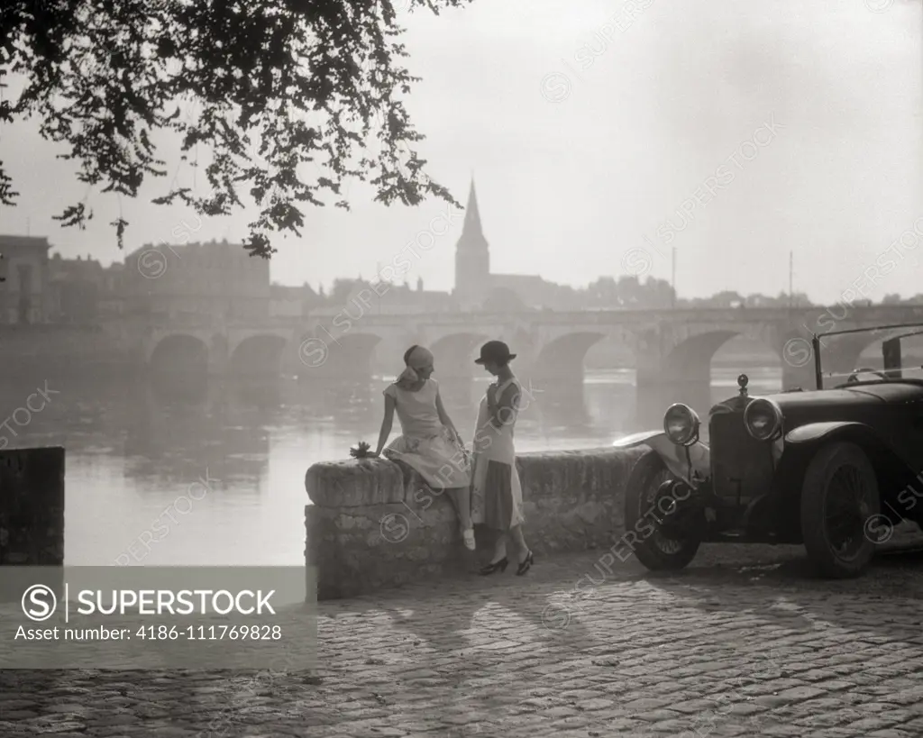 1920s TWO YOUNG WOMEN WITH ANTIQUE AUTOMOBILE SITTING STANDING TOGETHER TALKING BY STONE WALL LOIRE RIVER VALLEY FRANCE