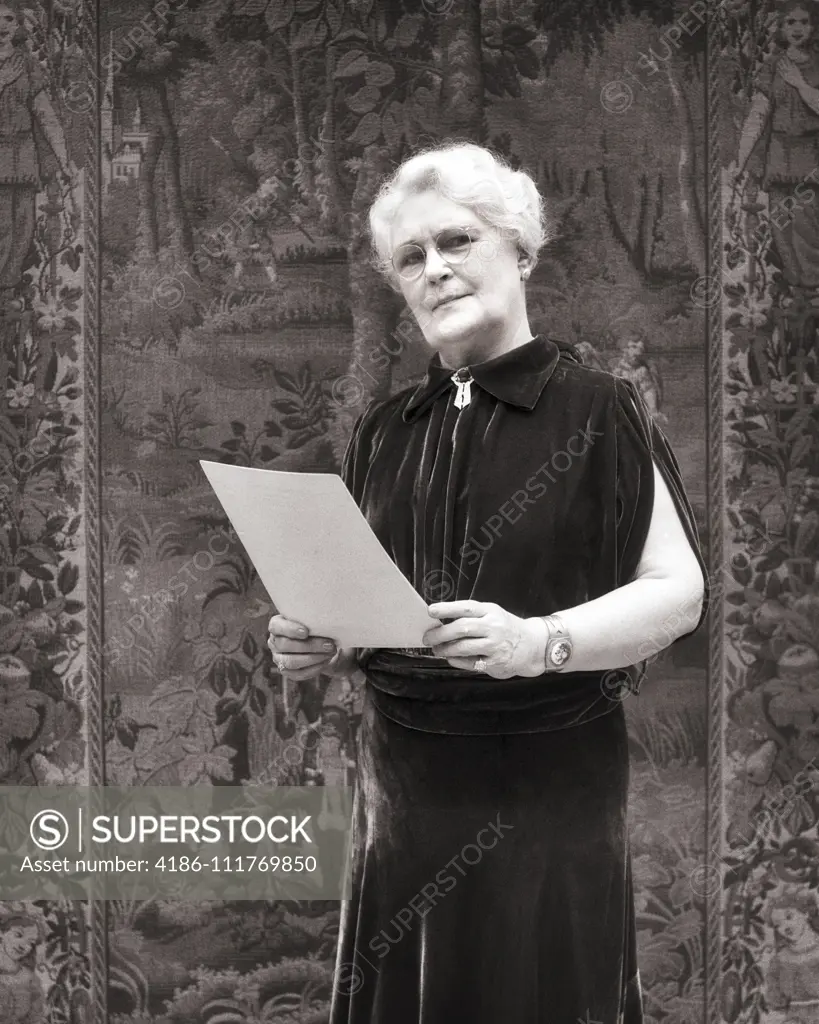 1930s elegant senior woman WEARING velvet dress holding paper GIVING A SPEECH STANDING BEFORE A TAPESTRY LOOKING AT CAMERA