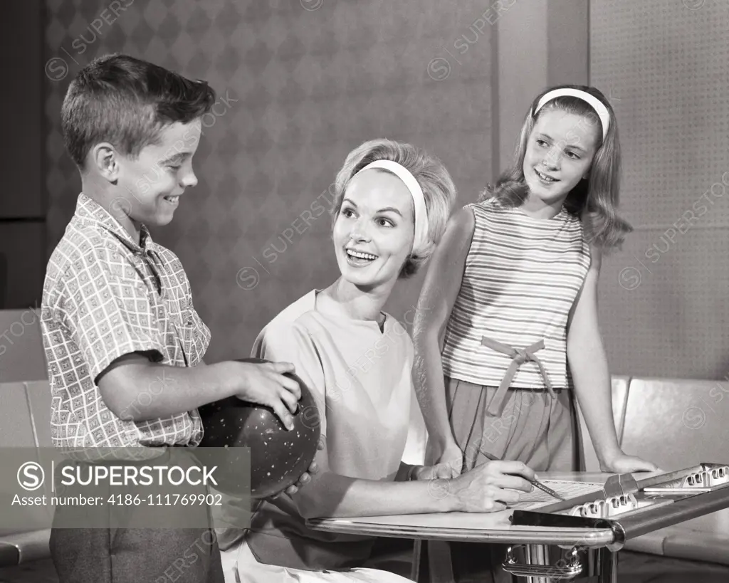 1960s Smiling mom keeping score at bowling alley with daughter and son who is holding a bowling ball