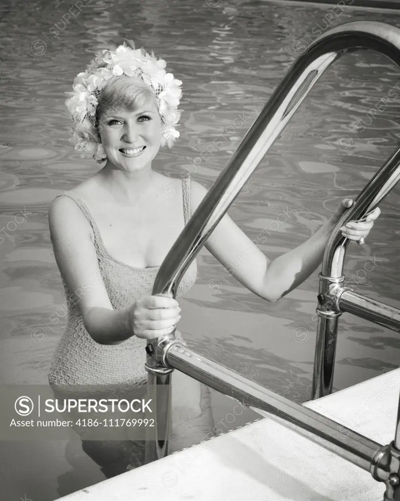 1960s BLOND WOMAN IN ONE PIECE BATHING SUIT AND FLOWER STYLE SWIM CAP CLIMBING OUT OF SWIMMING POOL SMILING LOOKING AT CAMERA