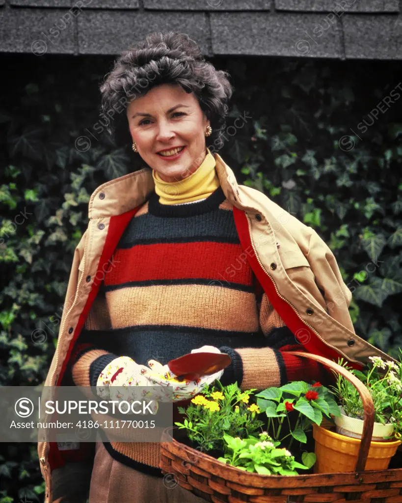 1980s SMILING WOMAN WEARING SWEATER JACKET YELLOW TURTLENECK GARDENING GLOVES HOLDING BASKET OF PLANTINGS LOOKING AT CAMERA