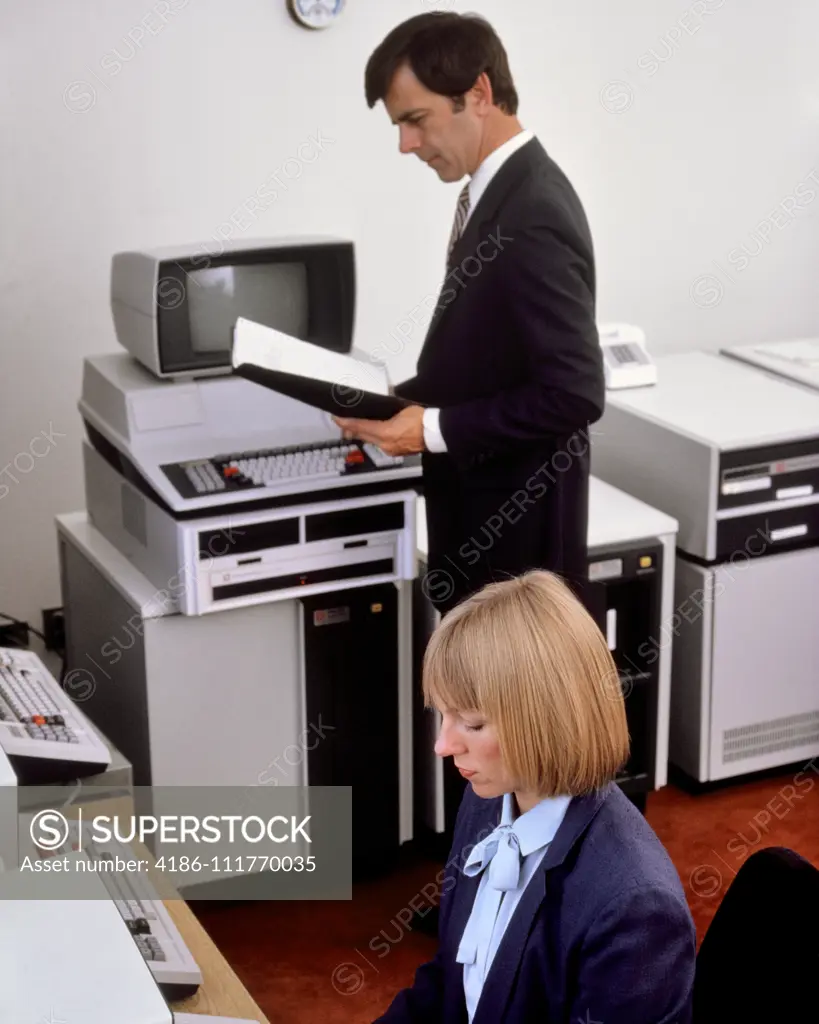 1980s OFFICE WITH EARLY COMPUTER HARDWARE WOMAN SEATED AT TERMINAL KEYBOARD MAN STANDING READING MANUAL