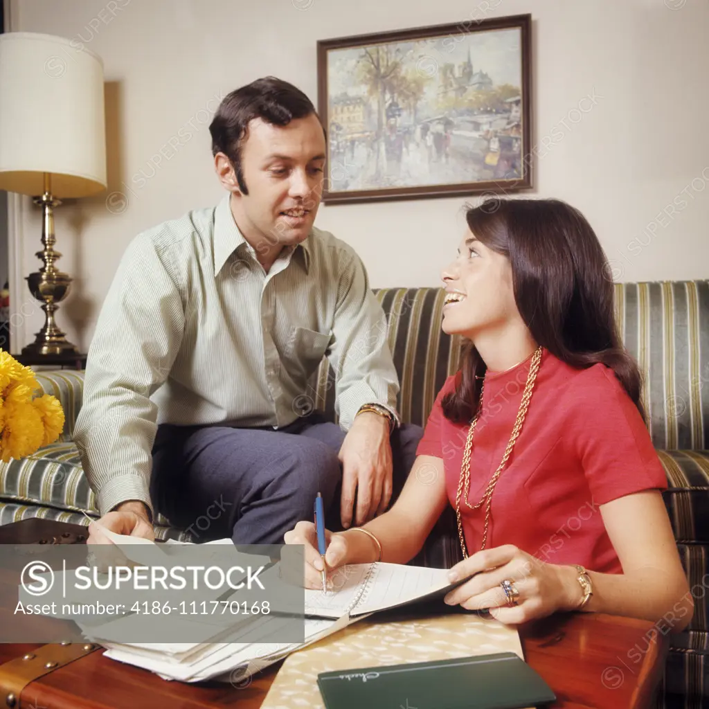 1970s SMILING COUPLE MAN WOMAN HUSBAND WIFE SITTING ON STRIPED COUCH REVIEWING FINANCES AND BILLS BALANCING CHECKBOOK CALENDAR