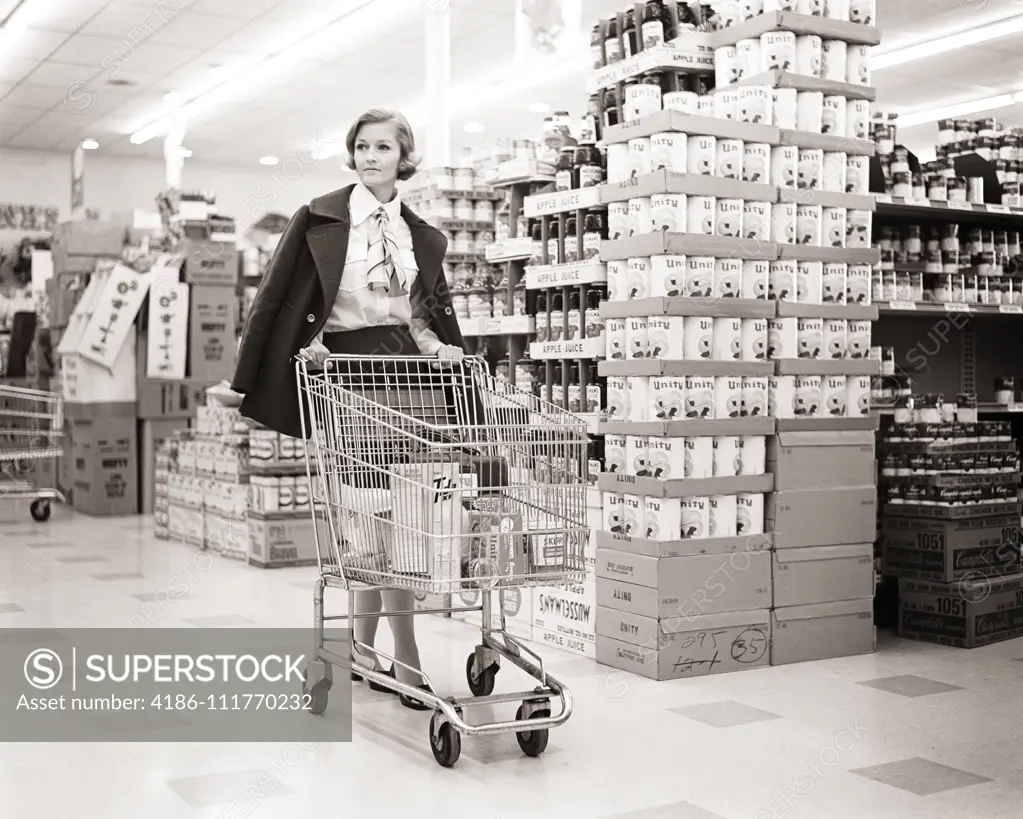 1960s 1970s WOMAN COAT DRAPED ON SHOULDERS PUSHING SHOPPING CART IN GROCERY STORE SUPERMARKET