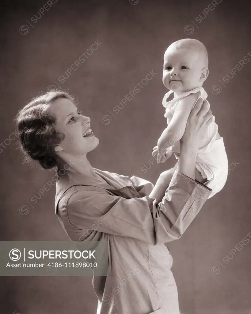 1930s SMILING MOTHER LIFTING UP BALD BABY BOY WITH FUNNY FACIAL EXPRESSION WEARING CLOTH DIAPER