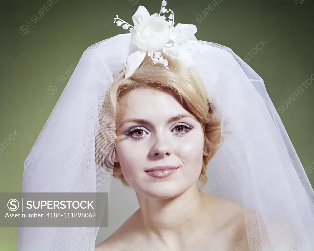 1960s PORTRAIT OF BLONDE BRIDE LOOKING AT CAMERA  WEARING NET VEIL WITH WHITE FLORAL ACCESSORY