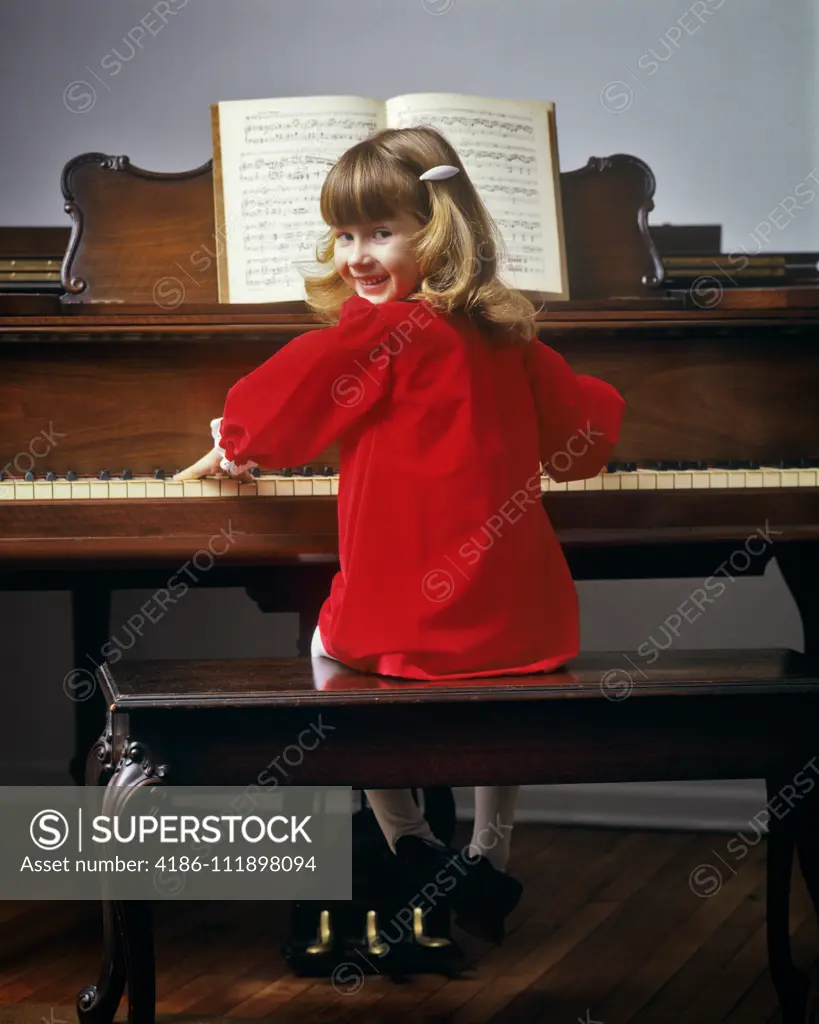 1980s CUTE SMILING YOUNG BLOND GIRL WEARING READ DRESS SITTING PLAYING PIANO TURNING HEAD OVER SHOULDER LOOKING AT CAMERA