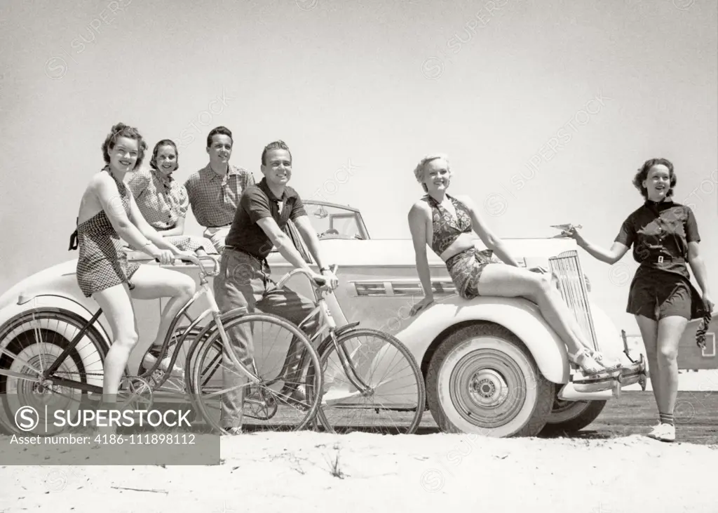 1930s GROUP OF MEN AND WOMEN WEARING BATHING SUITS CASUAL CLOTHES ON BICYCLES IN A CAR ON BEACH ALL LOOKING AT CAMERA SMILING