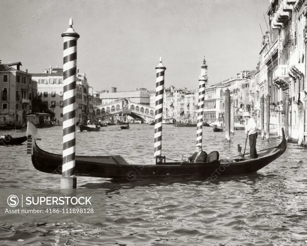 1950s ANONYMOUS BUSINESSMAN RIDING IN GONDOLA ROWING BOAT ON THE GRAND CANAL THE RIALTO BRIDGE IN BACKGROUND VENICE ITALY