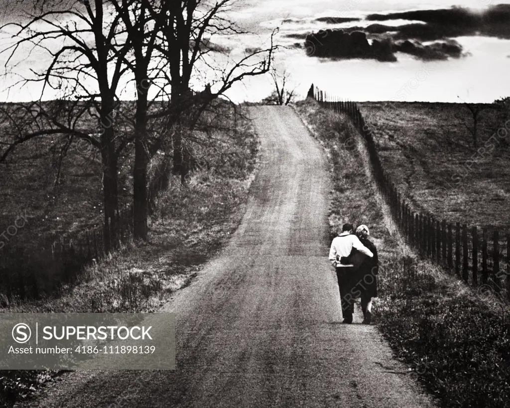 1960s BACK VIEW MATURE COUPLE ARM IN ARM WALKING DOWN LONELY ROAD BARREN AUTUMN LANDSCAPE WITH OMINOUS CLOUDS ON THE HORIZON