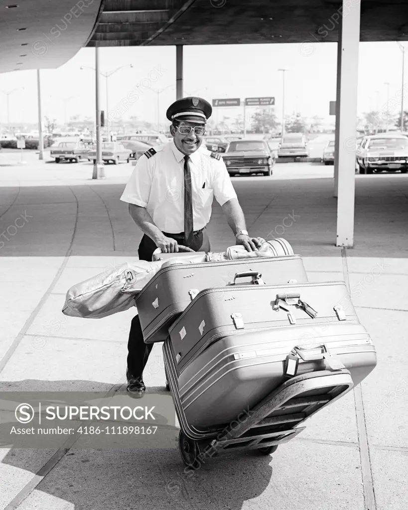 1960s SMILING AFRICAN-AMERICAN MAN AIRPORT PORTER PUSHING CART FULL OF LUGGAGE AT CURBSIDE