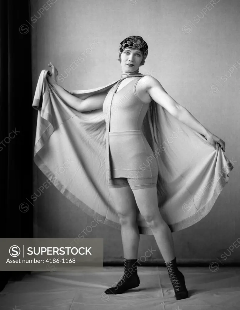 1920S Woman In Old-Fashioned Bathing Suit & Cap Holding Out Large Cape Tied Around Neck Looking At Camera