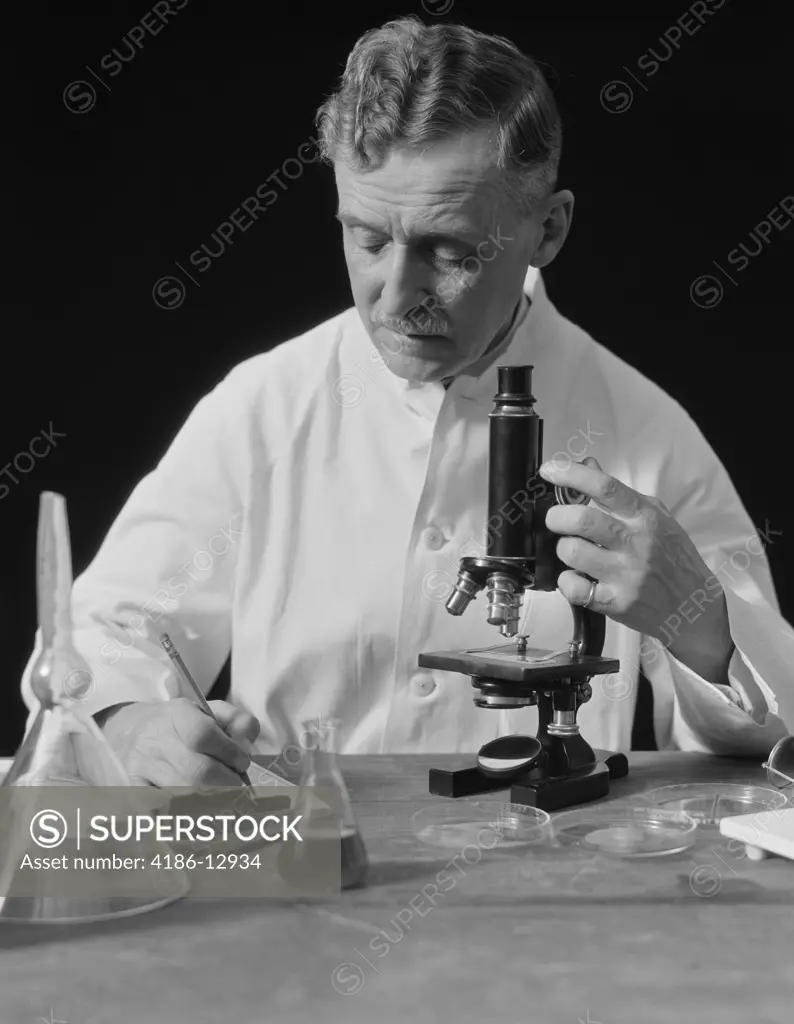 1920S 1930S Male Scientist Wearing White Lab Coat Writing Data On Clip Board Holding Focus Control Of Microscope