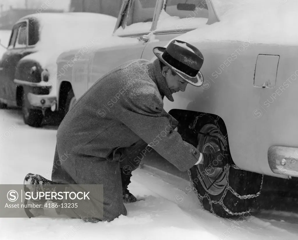 1950S Man In Coat & Hat Kneeling Down In Snow To Adjust Chains On Tires