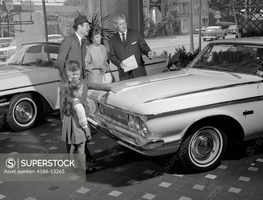 1960S Family Of 4 In Auto Showroom With Salesman Looking At White Plymouth