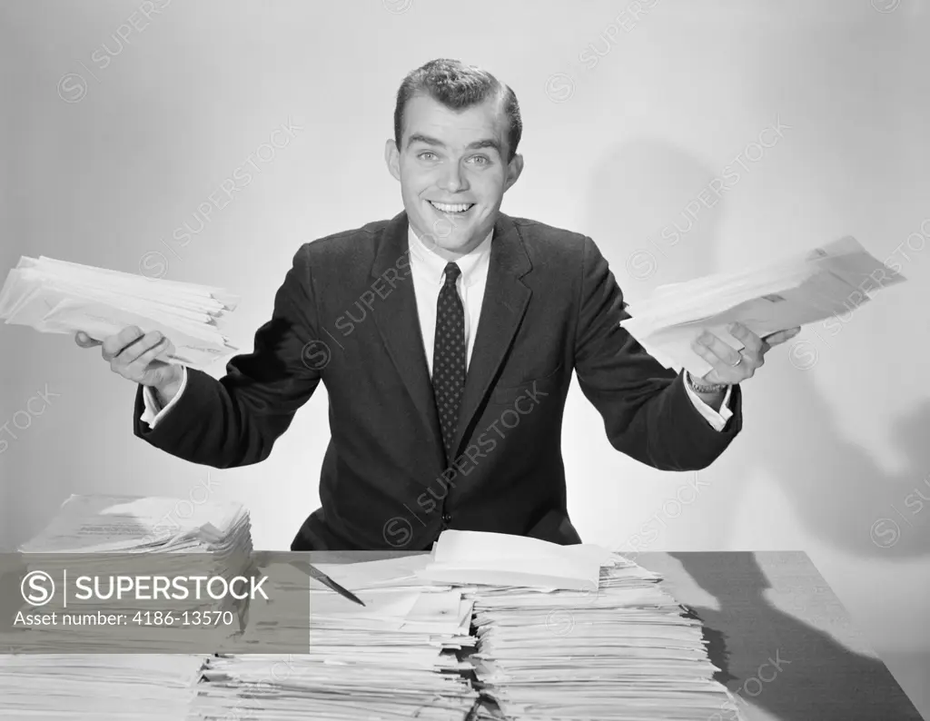 1950S Amused Smiling Businessman Holding Papers From The Piles On His Desk