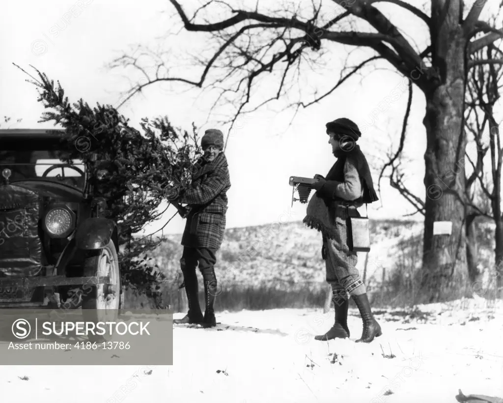 1920S Man Loading Christmas Tree Into Car With Woman Holding Camera Watching Him