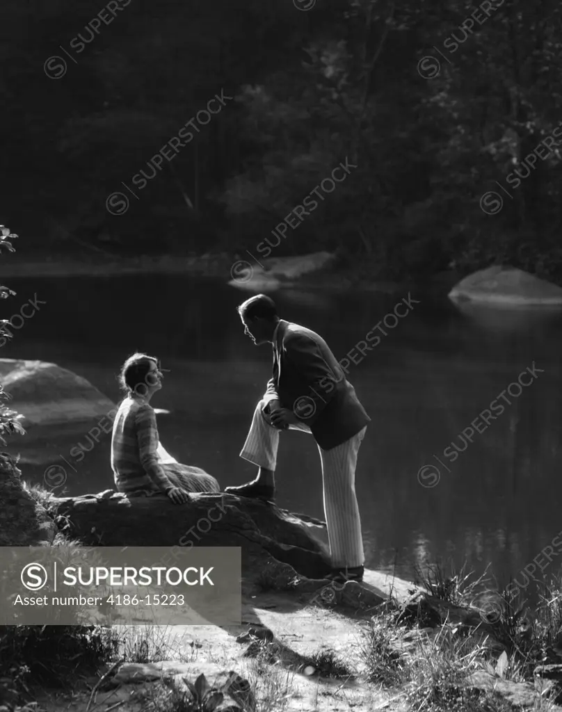 1920S 1930S Woman Sitting On Rock Lakeside Talking To Man Standing With Foot On Rock Leaning On Knee Listening