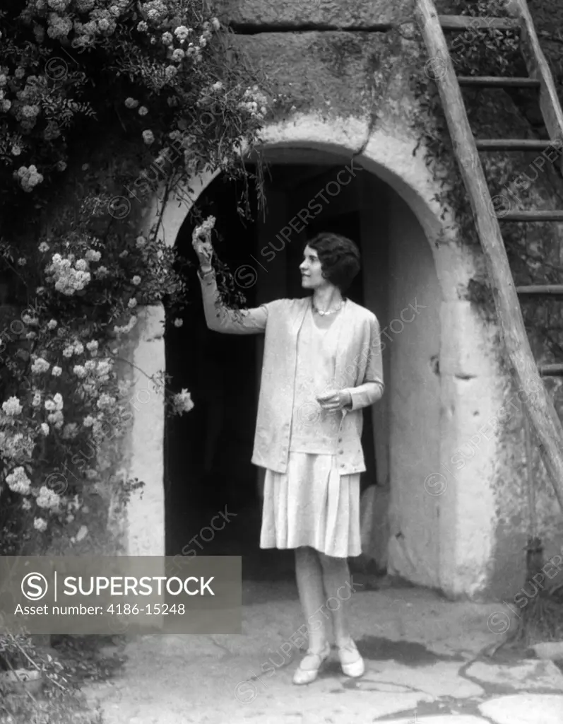 1930S Woman In Flapper Outfit Standing In Front Of Whitewashed Archway With Flowers Picking Bud Brittany France