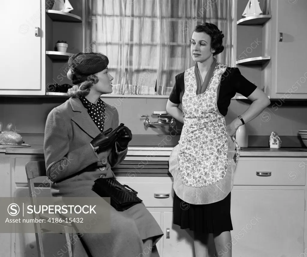 1930S 1940S Two Women Sitting In Kitchen Talking One Housewife Homemaker In Apron One In Coat And Hat Visiting Together Gossip